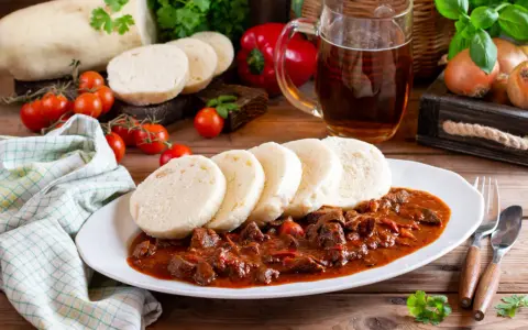 Gulasz z knedlikami, klasyczne czeskie potrawy, na białym talerzu. Obok kufel piwa, pomidorki i papryka.