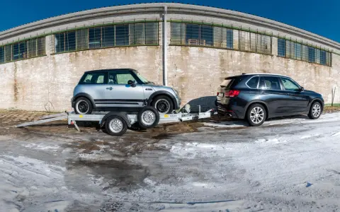 Srebrny Mini Cooper na lawecie, przygotowany do transportu. Obok stoi ciemny SUV. Mocowanie auta na lawecie jest kluczowe dla bezpiecznego przewozu.