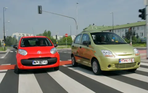 Czerwony i zielony samochód na pasach. Kierowca zielonego auta wykonuje manewr wyprzedzania czerwonego pojazdu.