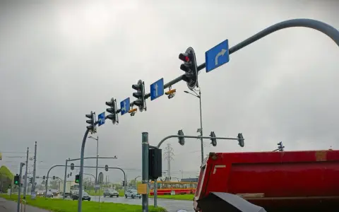 Na skrzyżowaniu wiszą sygnalizatory świetlne i znaki kierunkowe. Widać żółty tramwaj i czerwony wywrotkę. Gdzie są kamery rejestrujące przejazd na czerwonym świetle?