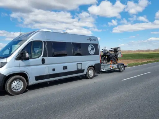 Srebrny kamper z motocyklem na przyczepie jedzie drogą. Przygoda w Szwecji kamperem dopiero się zaczyna.