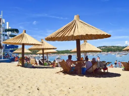 Słoneczna plaża na wyspie Rab, z ludźmi odpoczywającymi pod strzechami, w tle widać wodę z łodziami i zjeżdżalnie wodne.