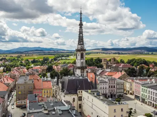 Urokliwe miasto na trasie Wrocław, z dominującą wieżą ratuszową i kolorowymi kamienicami, otoczone zielonymi polami i wzgórzami.