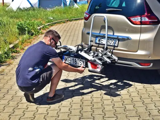 Mężczyzna montuje trzecią tablicę rejestracyjną WE 180SX na bagażniku rowerowym przy samochodzie.