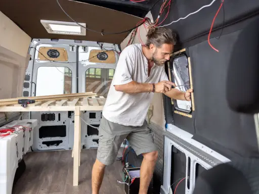 Mężczyzna buduje kamper z osobówki, montując okno. Widać drewniane elementy łóżka i instalację elektryczną.