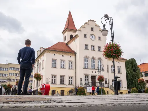 Mężczyzna stoi na rynku w Trzebnicy, podziwiając zabytkowy ratusz. Trasa spacerowa przez miasto prowadzi obok pięknych budynków.