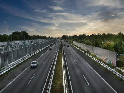 Szeroka autostrada z samochodami w oddali, otoczona zielenią i barierami dźwiękochłonnymi. Jazda autostradą pod pochmurnym niebem.
