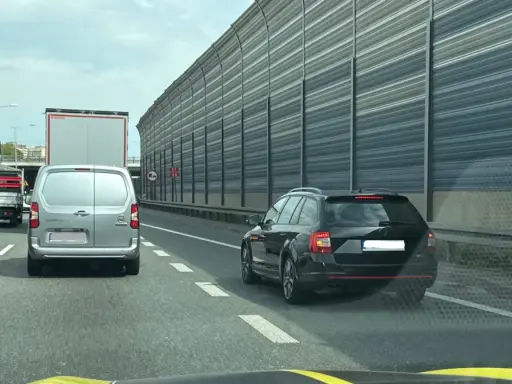 Czarny kombi jedzie pasem awaryjnym, ryzykując mandat. W tle inne pojazdy i ekrany akustyczne.