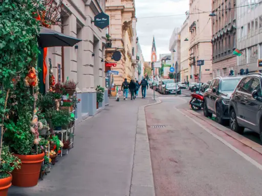 Wjazd do Wiednia samochodem osobowym. Ulica z kawiarniami, sklepami i zielenią, z zaparkowanymi autami i przechodniami.