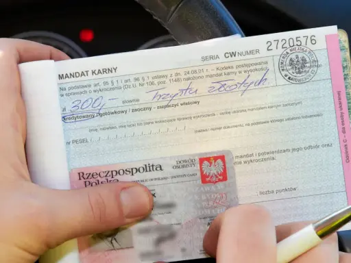 Ręka trzyma mandat karny na kwotę 300 zł. Obok dowód osobisty. Jak uniknąć mandatu? Uważaj na drodze!