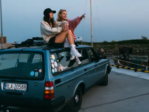 Dwie dziewczyny siedzą na dachu niebieskiego kombi, podziwiając widok na port. To idealny moment na podróż samochodem po Holandii.
