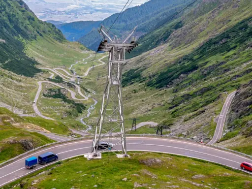 Kręte drogi w Rumunii wiją się przez zielone góry. Widać samochody i przyczepkę, a w oddali słup energetyczny.