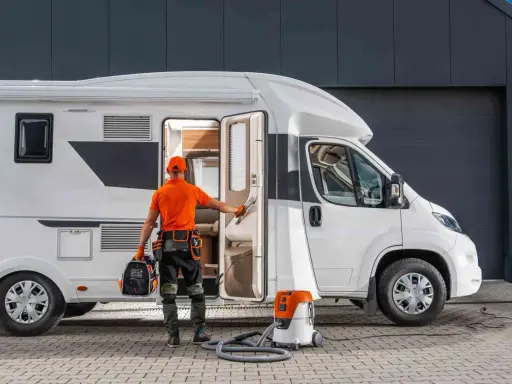 Fachowiec przygotowuje się do naprawy poszycia kampera z laminatu. W ręku trzyma narzędzia, obok stoi odkurzacz.