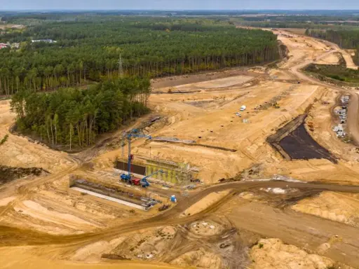 Ogromna budowa dróg w Polsce. Teren prac budowlanych z maszynami, dźwigami i przygotowywaną nawierzchnią, otoczony lasem.