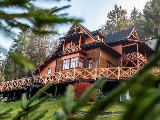 Drewniane schronisko turystyczne z tarasem i balustradą, otoczone drzewami.