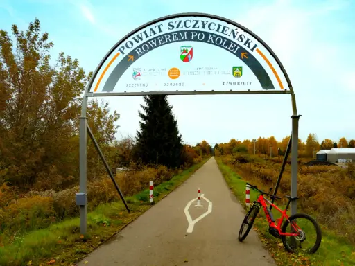 Pomarańczowy rower czeka na przygodę na trasie rowerowej w Szczytnie. Łuk nad drogą zaprasza do odkrywania powiatu.