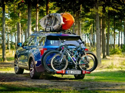 Niebieski SUV z dwoma rowerami na bagażniku i kajakami na dachu samochodu, gotowy na przygodę w lesie.