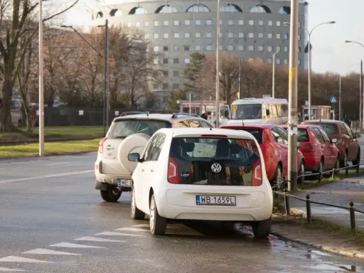 Biały Volkswagen up! zaparkowany tuż przy przejściu dla pieszych. W tle inne samochody i budynek.