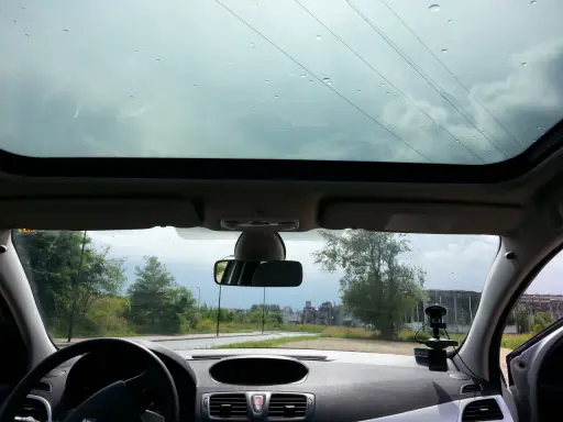 Widok z wnętrza samochodu z panoramicznym dachem, przez który widać niebo i drzewa. Krople deszczu na szybie.