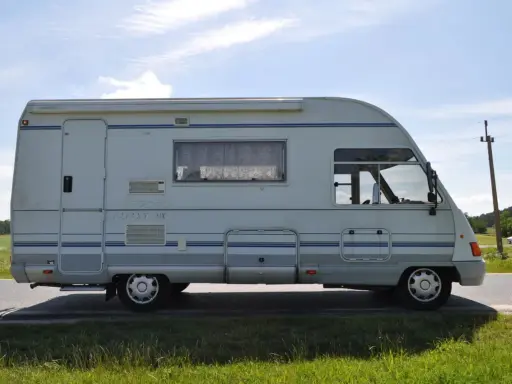 Biały ford galaxy camper z niebieskimi pasami stoi na poboczu drogi. W tle zielone pola i błękitne niebo.