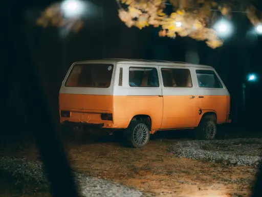 Pomarańczowo-biały kamper na nocnym parkingu, pod drzewami z lampkami. Czy można spać w kamperze na parkingu? Tak, to idealne miejsce na nocleg.