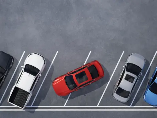 Czerwony samochód zajmuje dwa miejsca parkingowe, blokując sąsiednie pojazdy. Obok stoją czarny, biały pickup, srebrny i niebieski.