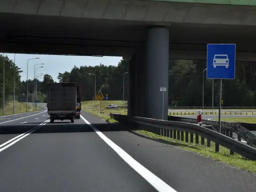 Droga jednojezdniowa pod wiaduktem, z ciężarówką i znakiem informującym o pasie ruchu dla samochodów.