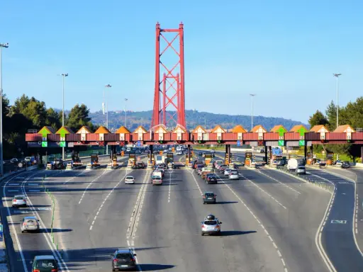 Ruchliwa autostrada w Portugalii z bramkami poboru opłat i charakterystycznym czerwonym mostem w tle.