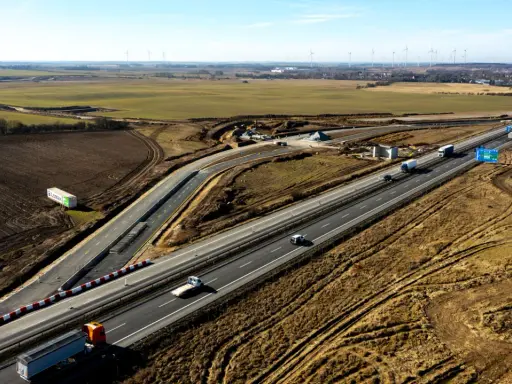 Rozbudowa autostrady A6 w otoczeniu pól i farm wiatrowych. Ruch samochodowy na ukończonych odcinkach, trwają prace budowlane.