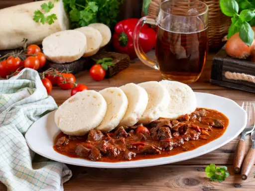 Gulasz z knedlikami, klasyczne czeskie potrawy, na białym talerzu. Obok kufel piwa, pomidorki i papryka.