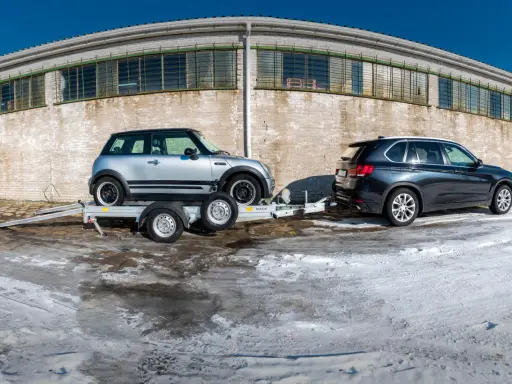Srebrny Mini Cooper na lawecie, przygotowany do transportu. Obok stoi ciemny SUV. Mocowanie auta na lawecie jest kluczowe dla bezpiecznego przewozu.