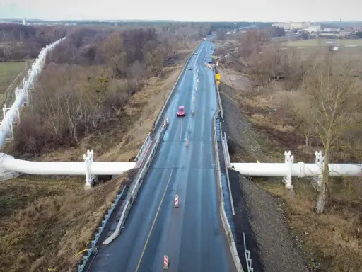 Droga krajowa 91 z wielkim białym rurociągiem biegnącym wzdłuż niej, otoczona drzewami i polami.