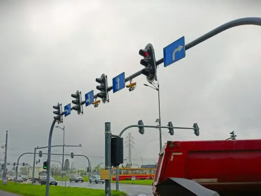Na skrzyżowaniu wiszą sygnalizatory świetlne i znaki kierunkowe. Widać żółty tramwaj i czerwony wywrotkę. Gdzie są kamery rejestrujące przejazd na czerwonym świetle?