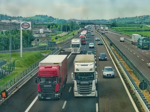 Ciężarówki na autostradzie, jedna czerwona Scania wyprzedza białą. W tle inne pojazdy i krajobraz z pagórkami.
