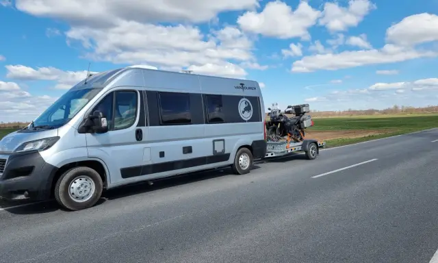 Srebrny kamper z motocyklem na przyczepie jedzie drogą. Przygoda w Szwecji kamperem dopiero się zaczyna.