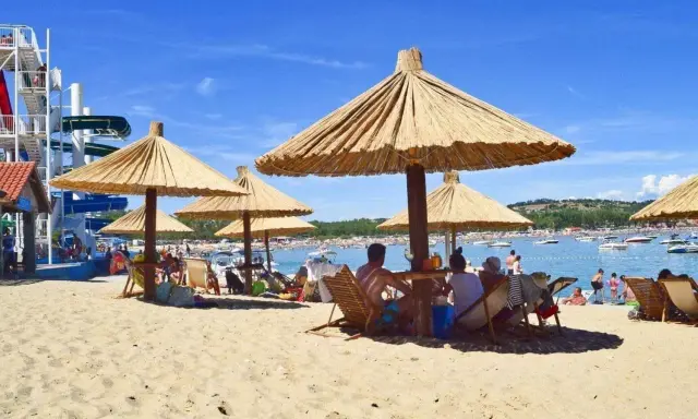 Słoneczna plaża na wyspie Rab, z ludźmi odpoczywającymi pod strzechami, w tle widać wodę z łodziami i zjeżdżalnie wodne.