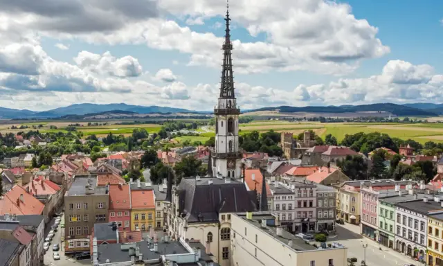 Urokliwe miasto na trasie Wrocław, z dominującą wieżą ratuszową i kolorowymi kamienicami, otoczone zielonymi polami i wzgórzami.
