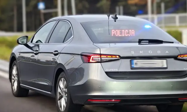 Szary, nieoznakowany radiowóz Skoda Superb z włączonymi niebieskimi światłami i napisem "POLICJA" na tylnej szybie.