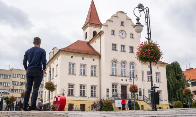 Mężczyzna stoi na rynku w Trzebnicy, podziwiając zabytkowy ratusz. Trasa spacerowa przez miasto prowadzi obok pięknych budynków.