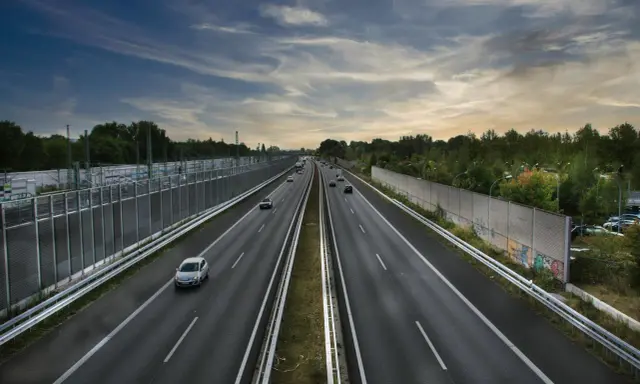 Szeroka autostrada z samochodami w oddali, otoczona zielenią i barierami dźwiękochłonnymi. Jazda autostradą pod pochmurnym niebem.