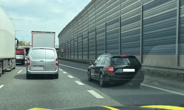 Czarny kombi jedzie pasem awaryjnym, ryzykując mandat. W tle inne pojazdy i ekrany akustyczne.