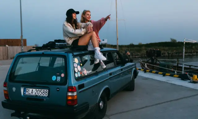 Dwie dziewczyny siedzą na dachu niebieskiego kombi, podziwiając widok na port. To idealny moment na podróż samochodem po Holandii.