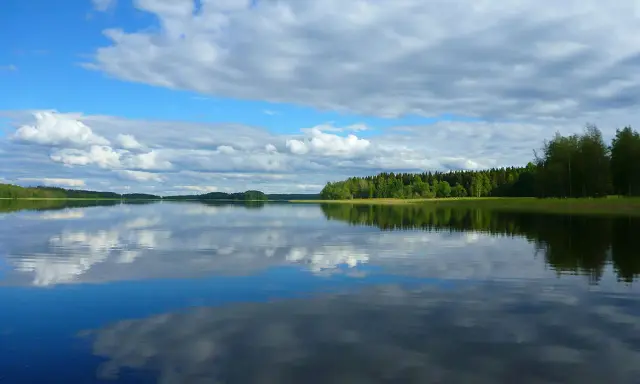 Spokojne jezioro na pojezierzu fińskim, odbijające chmury i zielone lasy.