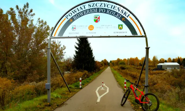 Pomarańczowy rower czeka na przygodę na trasie rowerowej w Szczytnie. Łuk nad drogą zaprasza do odkrywania powiatu.