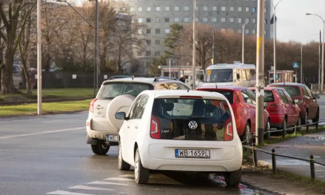Biały Volkswagen up! zaparkowany tuż przy przejściu dla pieszych. W tle inne samochody i budynek.
