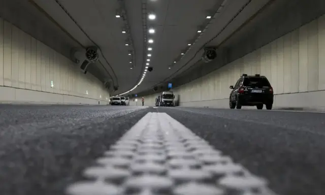 Wnętrze tunelu z samochodami i ciężarówką. Widać wyraźnie jaki odstep w tunelu między pojazdami, oświetlenie i wentylatory.
