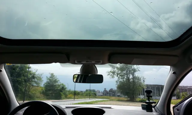 Widok z wnętrza samochodu z panoramicznym dachem, przez który widać niebo i drzewa. Krople deszczu na szybie.
