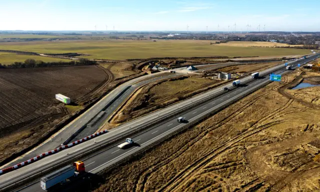 Rozbudowa autostrady A6 w otoczeniu pól i farm wiatrowych. Ruch samochodowy na ukończonych odcinkach, trwają prace budowlane.