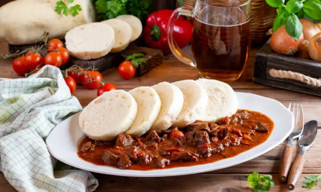 Gulasz z knedlikami, klasyczne czeskie potrawy, na białym talerzu. Obok kufel piwa, pomidorki i papryka.