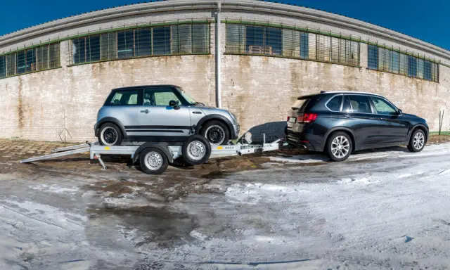 Srebrny Mini Cooper na lawecie, przygotowany do transportu. Obok stoi ciemny SUV. Mocowanie auta na lawecie jest kluczowe dla bezpiecznego przewozu.